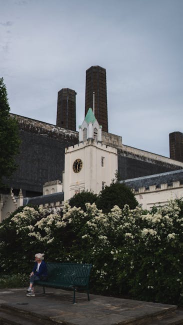 A riverside view featuring a garden scene where a well-maintained lawn with trimmed grass borders a paved pathway, bordered by neatly pruned hedges and a variety of plants. In the background, tall dec